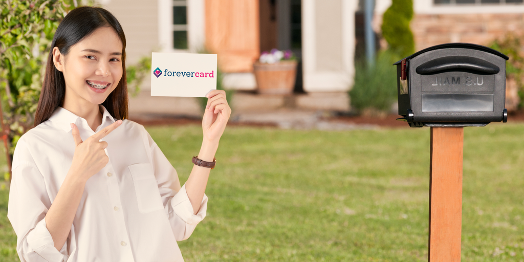 Smiling woman standing in front of a suburban house, holding and pointing to a ForeverCard-branded card near a black U.S. mailbox, symbolizing sending or receiving cards.