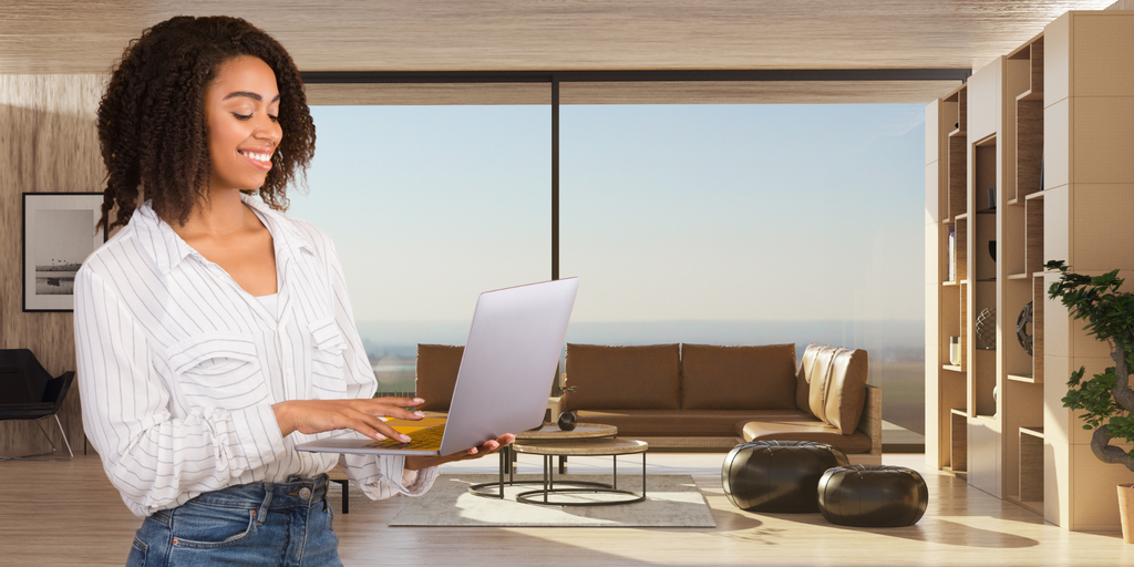 Smiling woman working on a laptop in a modern, sunlit living room with large windows overlooking a scenic view, surrounded by minimalist furniture and shelves.